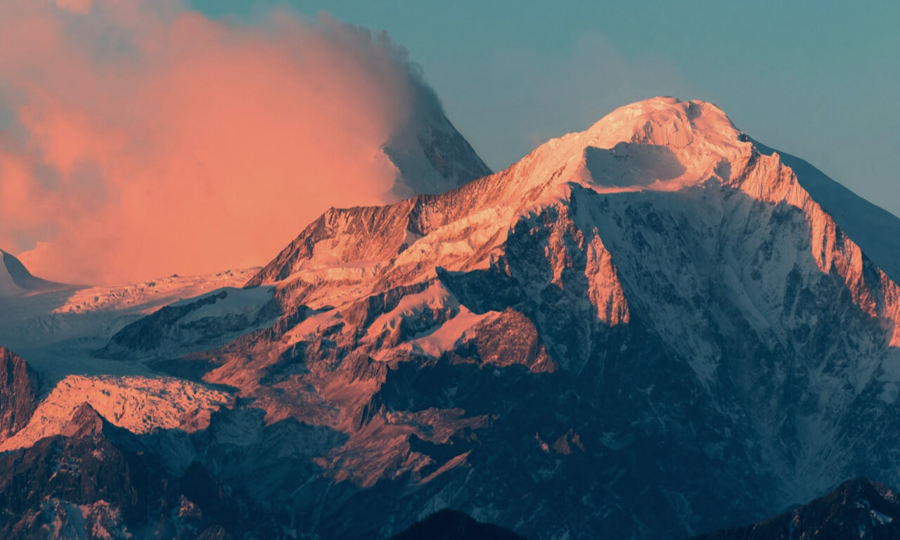 A pair of snowy mountain peaks, glowing pink from the early morning sunlight, against a teal blue sky