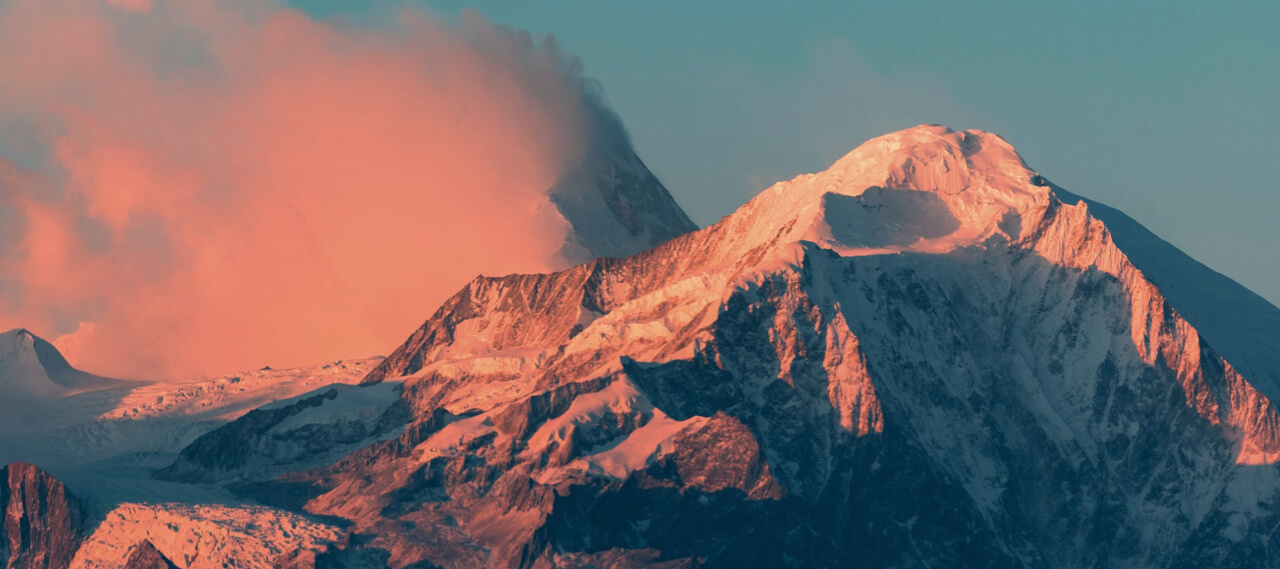 Close-in crop of two tall mountain peaks, one shrouded in blowing snow, both illuminated by the pink-orange light of sunrise
